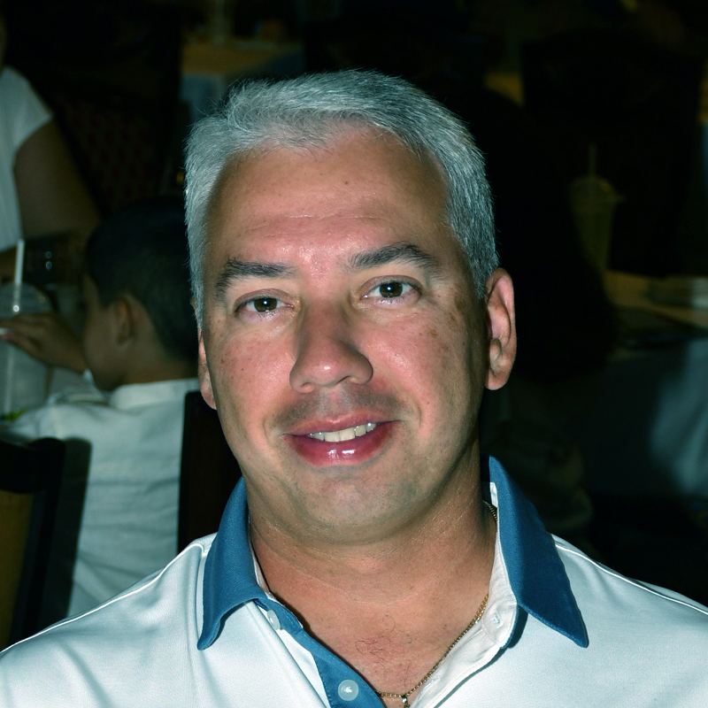 Foto grafía de Roberto con fondo negro, camisa blanca con cuello azul.